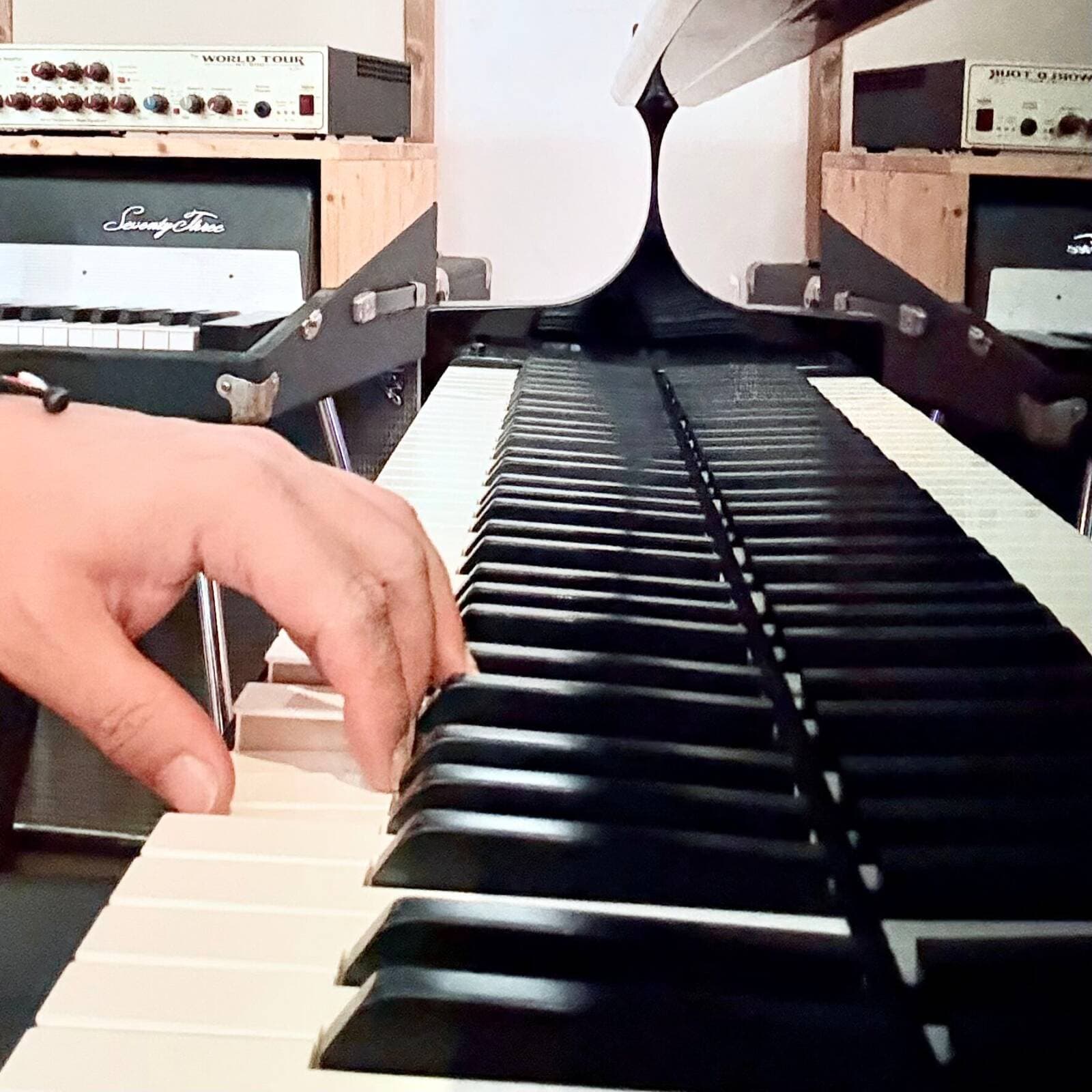 A hand playing the keys of the studio grand piano, with vintage keyboards and amplifiers behind it