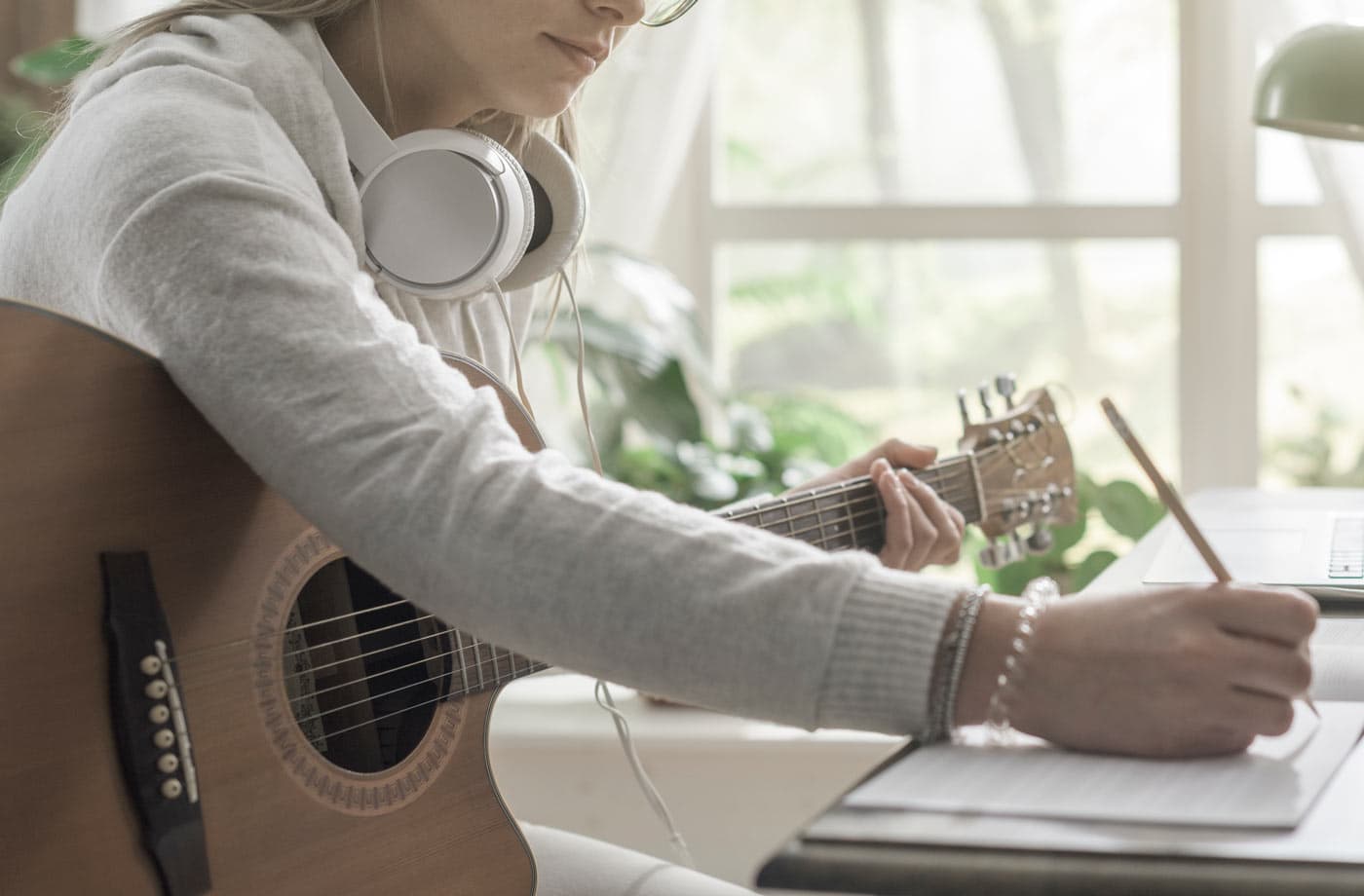 A songwriter with headphones around her neck playing an acoustic guitar while writing at a desk by a window